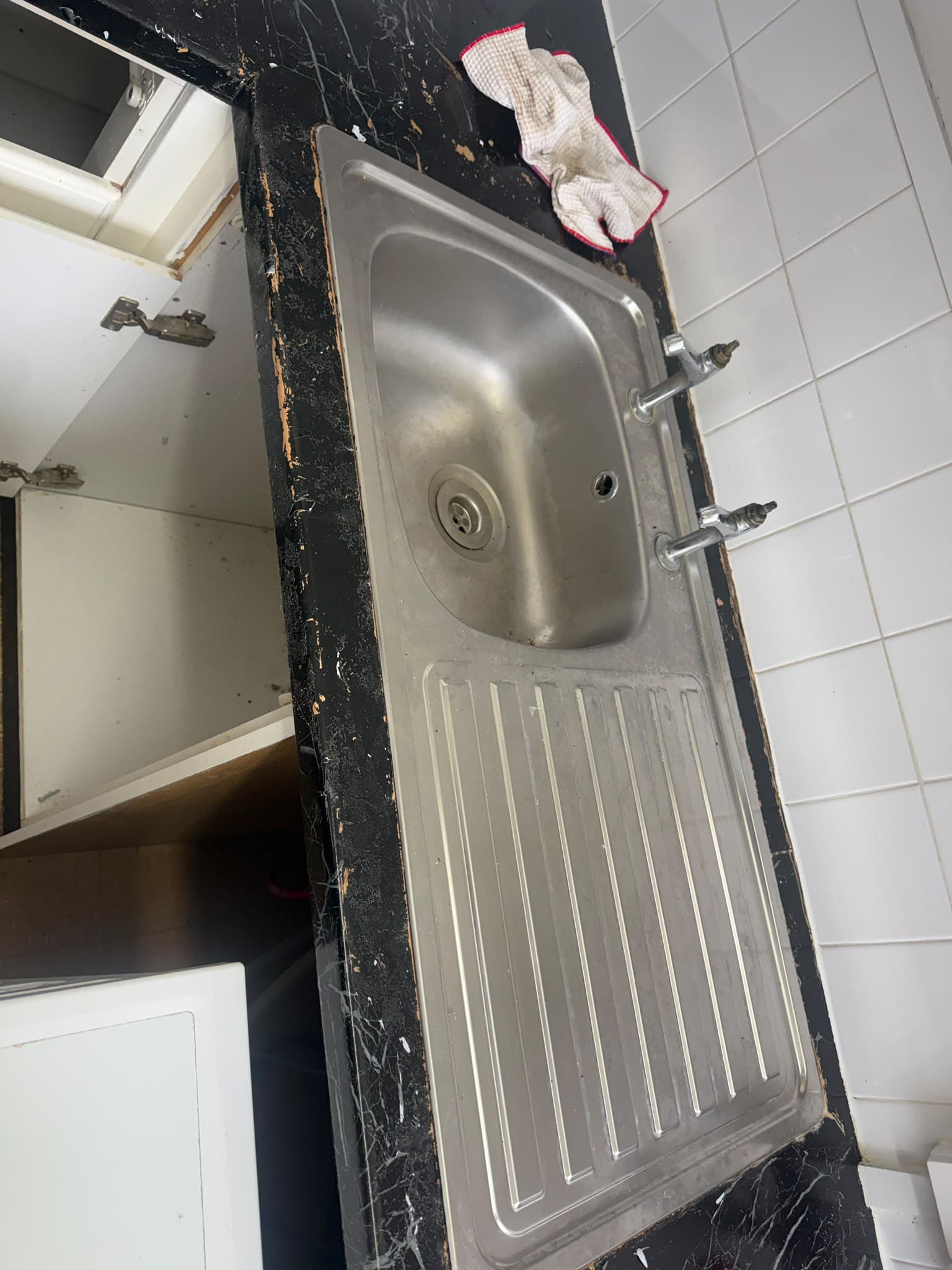 A stainless steel kitchen sink with taps removed, installed in a worn black countertop with a cloth resting on it.