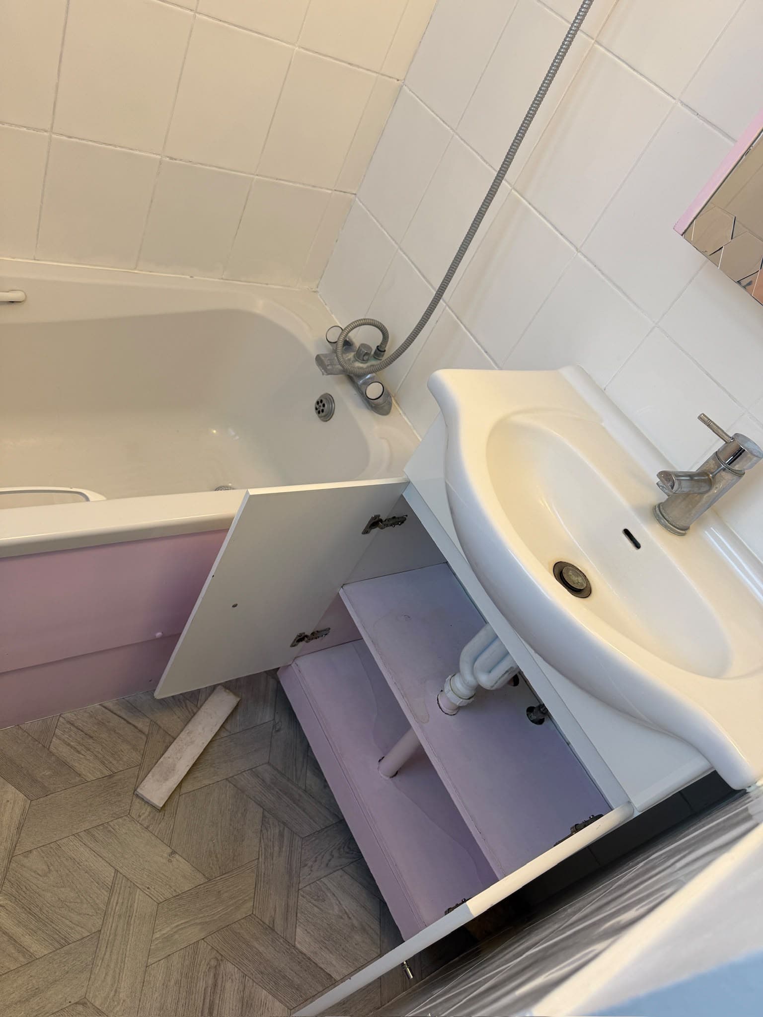 A bathroom with a tub and sink, showing an open cabinet panel revealing plumbing pipework underneath the sink.