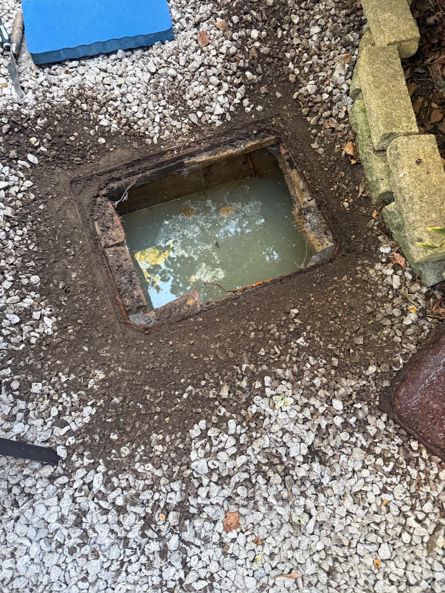 An open drain or inspection chamber filled with murky water surrounded by gravel and soil
