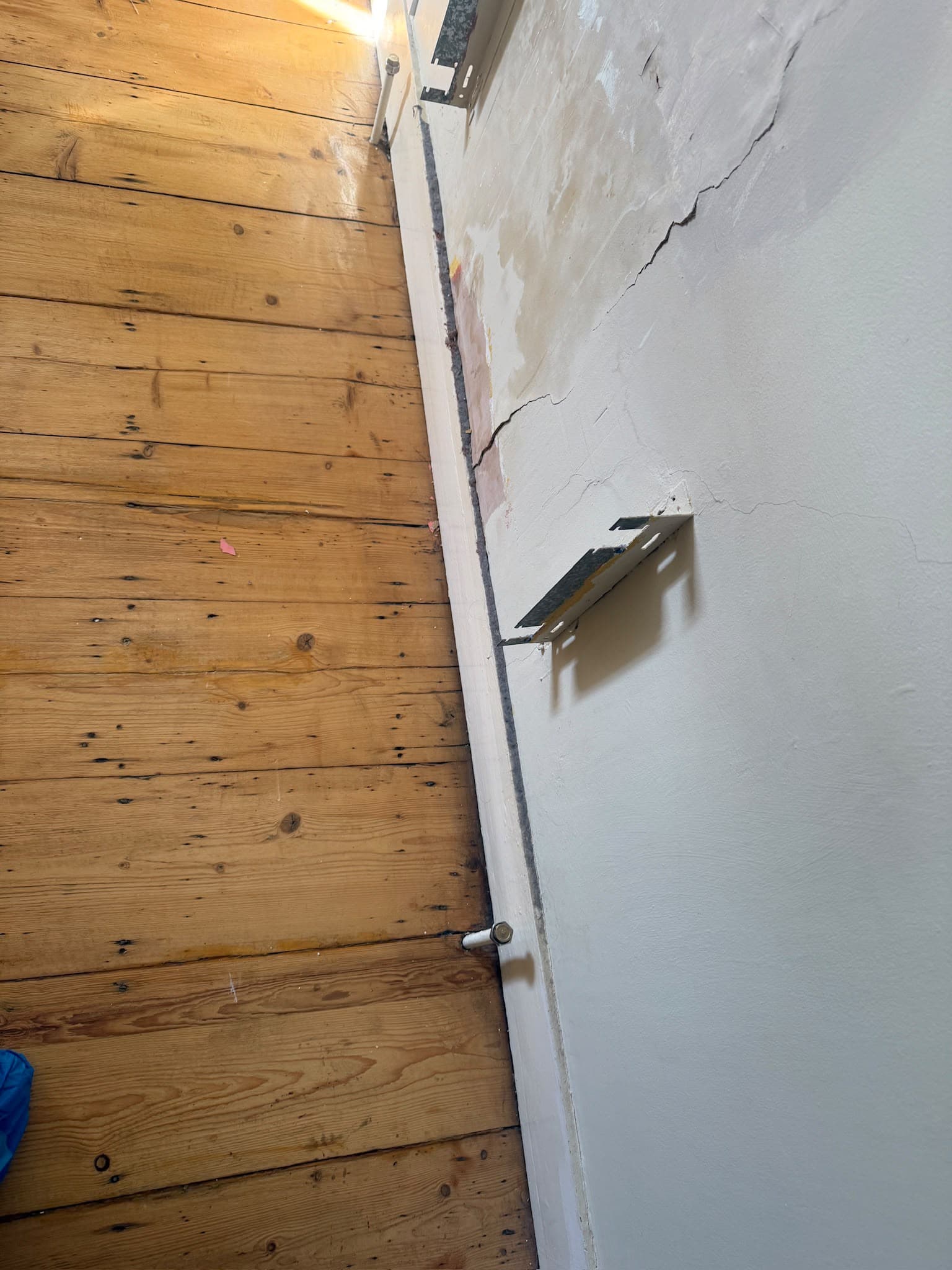 A wooden floor meeting a cracked plaster wall with exposed repair brackets and skirting board removed, showing a home renovation site